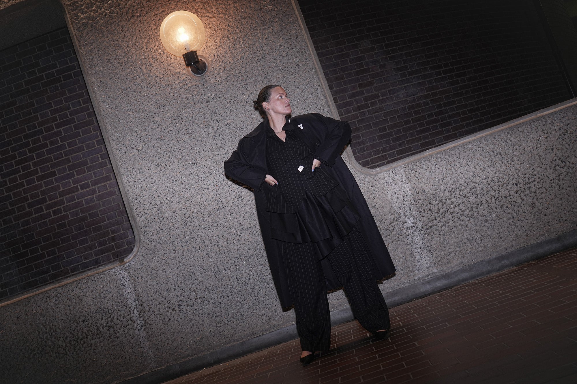 Person wearing a black coat and pinstripe pants standing against a textured wall with a light fixture.