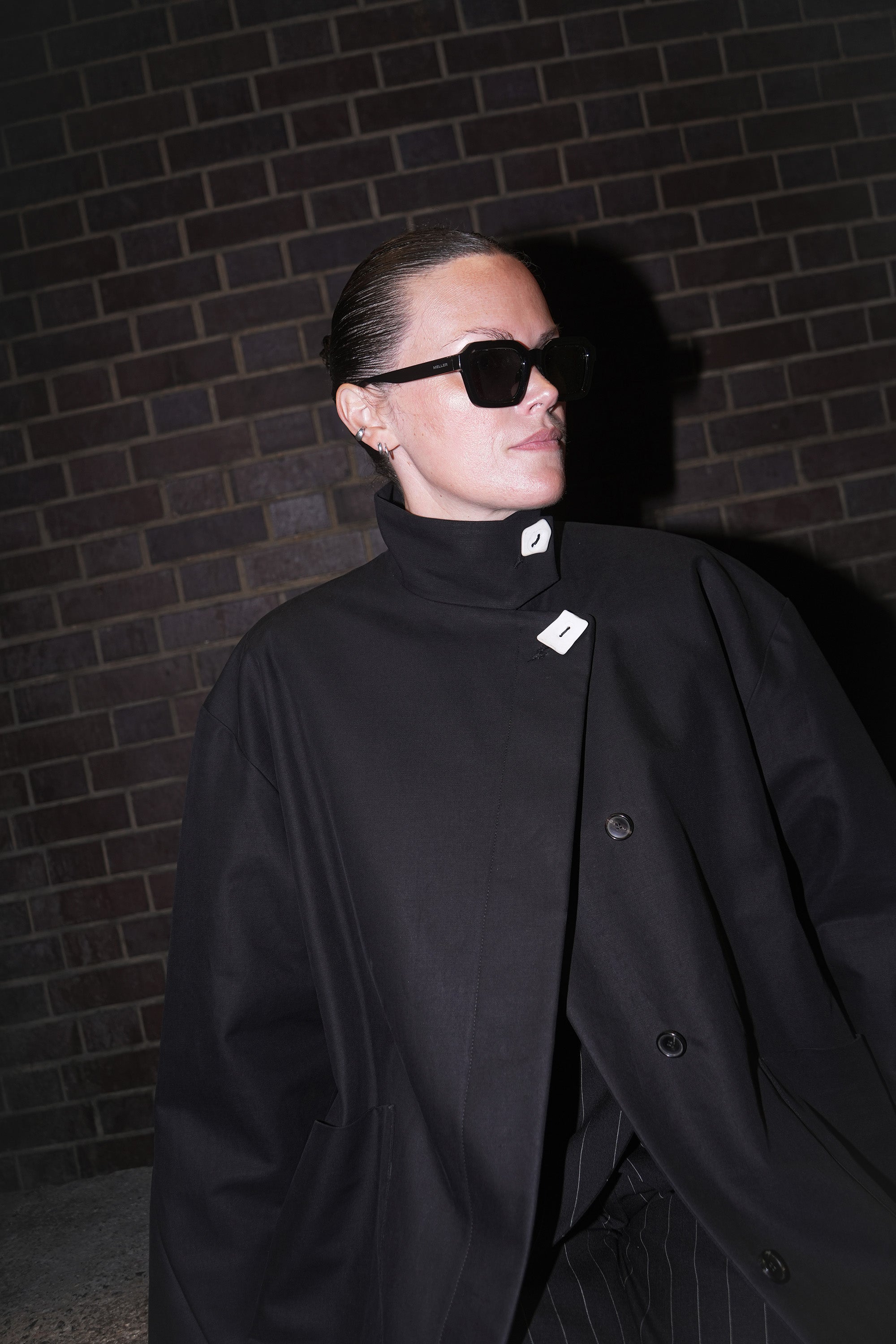 Person wearing the black 'Hilda' coat and sunglasses against a brick wall.