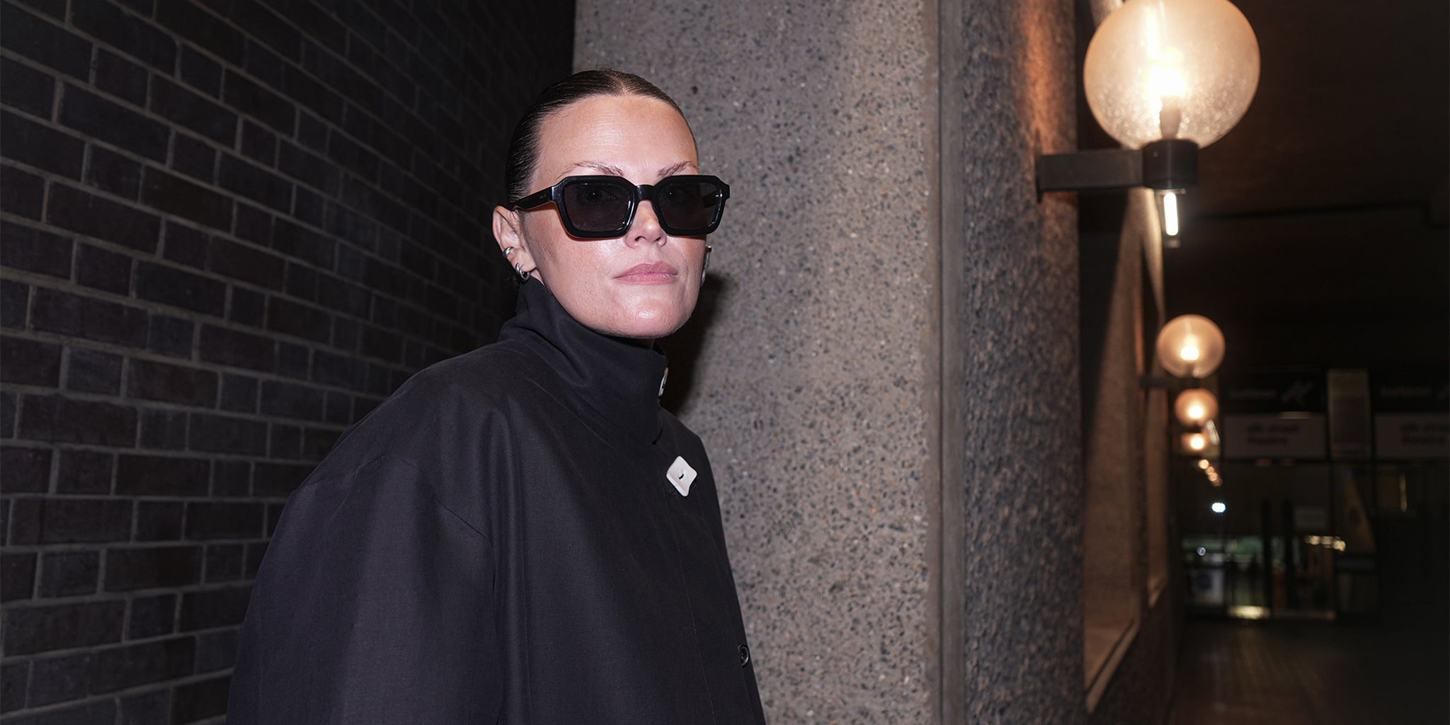 Person wearing a black outfit and sunglasses standing against a brick wall at night.